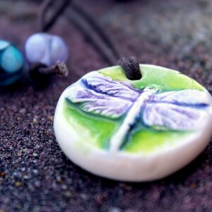 May include: A ceramic pendant necklace featuring a dragonfly design. The pendant is white with a green and purple dragonfly, and is strung on a dark brown cord. Two decorative beads in teal and lavender are also visible.