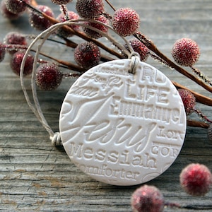 May include: A round, off-white ornament with the word "Jesus" prominently displayed, along with other religious terms. The ornament hangs from a twine loop and rests near a sprig of red berries.