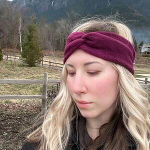 Puede incluir: Una diadema de forro polar burdeos oscuro con un diseño de nudo retorcido. La diadema está hecha de un material suave y texturizado. El fondo presenta una cordillera y un cielo nublado.