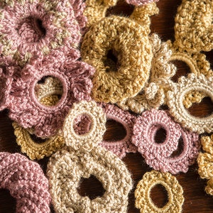 K&ouml;nnte beinhalten: Eine Sammlung geh&auml;kelter Blumen-Haargummis in Rosa und Beige. Die Haargummis sind mit verschiedenen Maschen und Texturen gearbeitet.