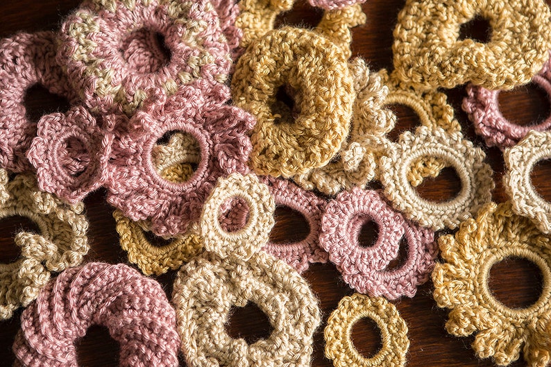 K&ouml;nnte beinhalten: Eine Sammlung geh&auml;kelter Blumen-Haargummis in Rosa und Beige. Die Haargummis sind mit verschiedenen Maschen und Texturen gearbeitet.