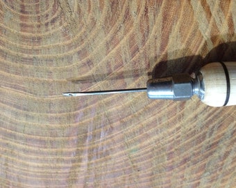 Puede incluir: Primer plano de un punzón de cuero con mango de madera y una aguja de metal. El punzón está sobre una superficie de madera.