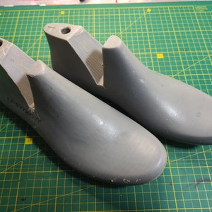 Puede incluir: Dos hormas de zapatos grises, utilizadas para dar forma a los zapatos, están sobre una estera de corte verde. Las hormas están hechas de plástico liso y duro.