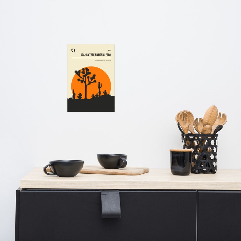 May include: A minimalist poster with a beige background featuring a black silhouette of a Joshua Tree National Park landscape with a large orange sun. The text "Joshua Tree National Park" is printed in black at the top of the poster.