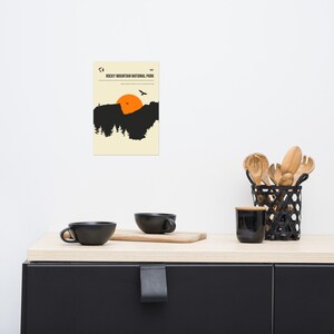 Op de afbeelding: Een minimalistische poster met een zwart silhouet van een bergketen en een ondergaande zon. De tekst "Rocky Mountain National Park" is bovenaan de poster in zwart gedrukt.