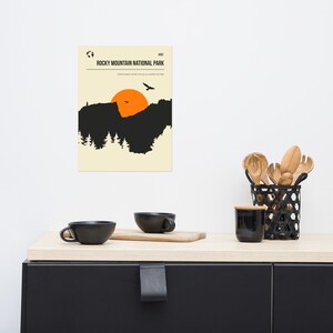 Op de afbeelding: Een minimalistische poster met een zwart silhouet van een bergketen en een feloranje zon die erachter ondergaat. De tekst "VISIT ROCKY MOUNTAIN NATIONAL PARK" staat bovenaan de poster.