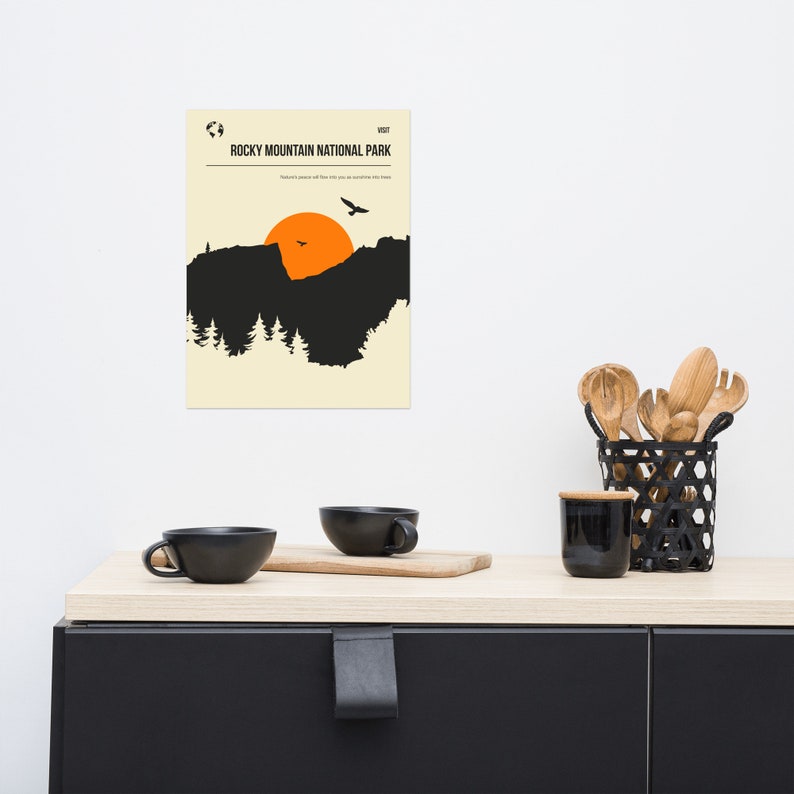 Op de afbeelding: Een minimalistische poster met een zwart silhouet van een bergketen en een feloranje zon die erachter ondergaat. De tekst "VISIT ROCKY MOUNTAIN NATIONAL PARK" staat bovenaan de poster.
