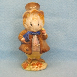 May include: A ceramic figurine of a mouse dressed in a brown coat, blue bow tie, and top hat. The mouse is holding a small yellow object and a blue book. The base is a light brown with red and orange accents.