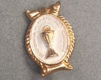 May include: Gold-toned oval pin with a white center featuring a chalice and the text "REMEMBERANCE OF MY FIRST HOLY COMMUNION". The pin has a rope-like border and decorative elements.