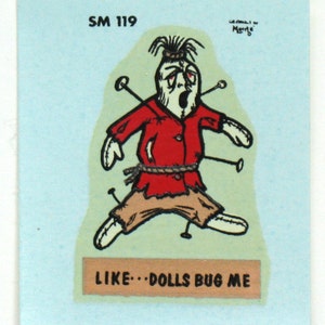 May include: A vintage cartoon illustration of a voodoo doll with red and brown clothing, pinned with needles. The text "LIKE...DOLLS BUG ME" is printed on a banner below the doll.
