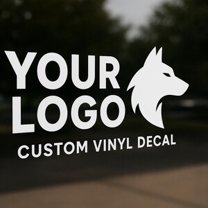 May include: White vinyl decal on glass with the text "YOUR LOGO" and "CUSTOM VINYL DECAL". A white wolf head graphic is to the right of the text. The background is blurred.
