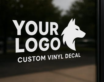 May include: White vinyl decal on glass with the text "YOUR LOGO" and "CUSTOM VINYL DECAL". A white wolf head graphic is to the right of the text. The background is blurred.