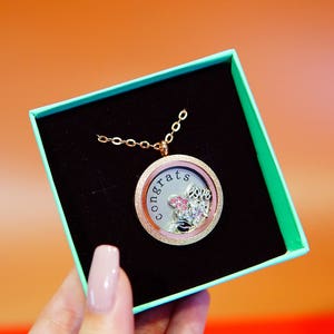 May include: A gold locket necklace with a pink and white charm that says "Congrats" and "Love". The locket is in a teal gift box with a black interior.