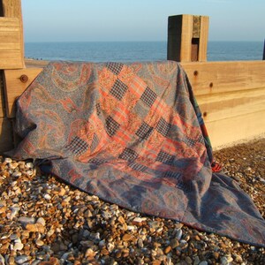 May include: A patchwork quilt with a paisley pattern and red and blue plaid squares is lying on a rocky beach near a wooden fence.