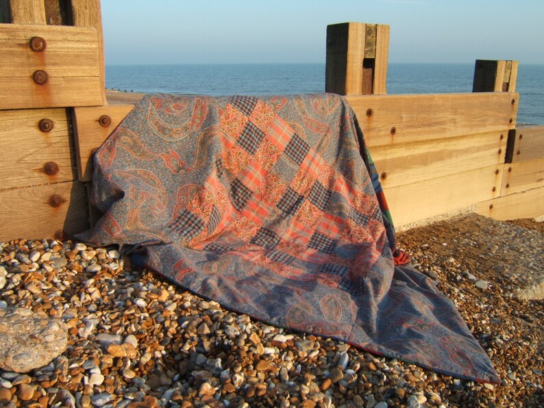 May include: A patchwork quilt with a paisley pattern and red and blue plaid squares is lying on a rocky beach near a wooden fence.