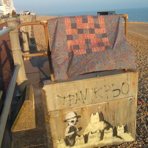 May include: A concrete wall with graffiti that says "Team K350". A stencil of a child building sandcastles with the word "Tesco" on each one is painted on the wall. The child is wearing a hat and sunglasses. A colorful quilt is draped over a metal railing in the background.