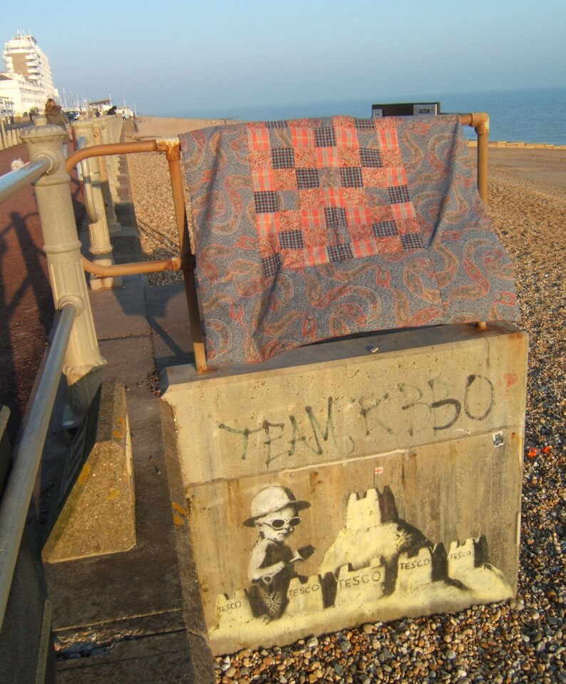 May include: A concrete wall with graffiti that says "Team K350". A stencil of a child building sandcastles with the word "Tesco" on each one is painted on the wall. The child is wearing a hat and sunglasses. A colorful quilt is draped over a metal railing in the background.