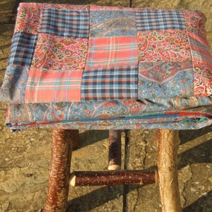 May include: A patchwork quilt made of blue, red, and white fabric is draped over a wooden stool with a natural bark finish. The stool has three legs and a simple design.