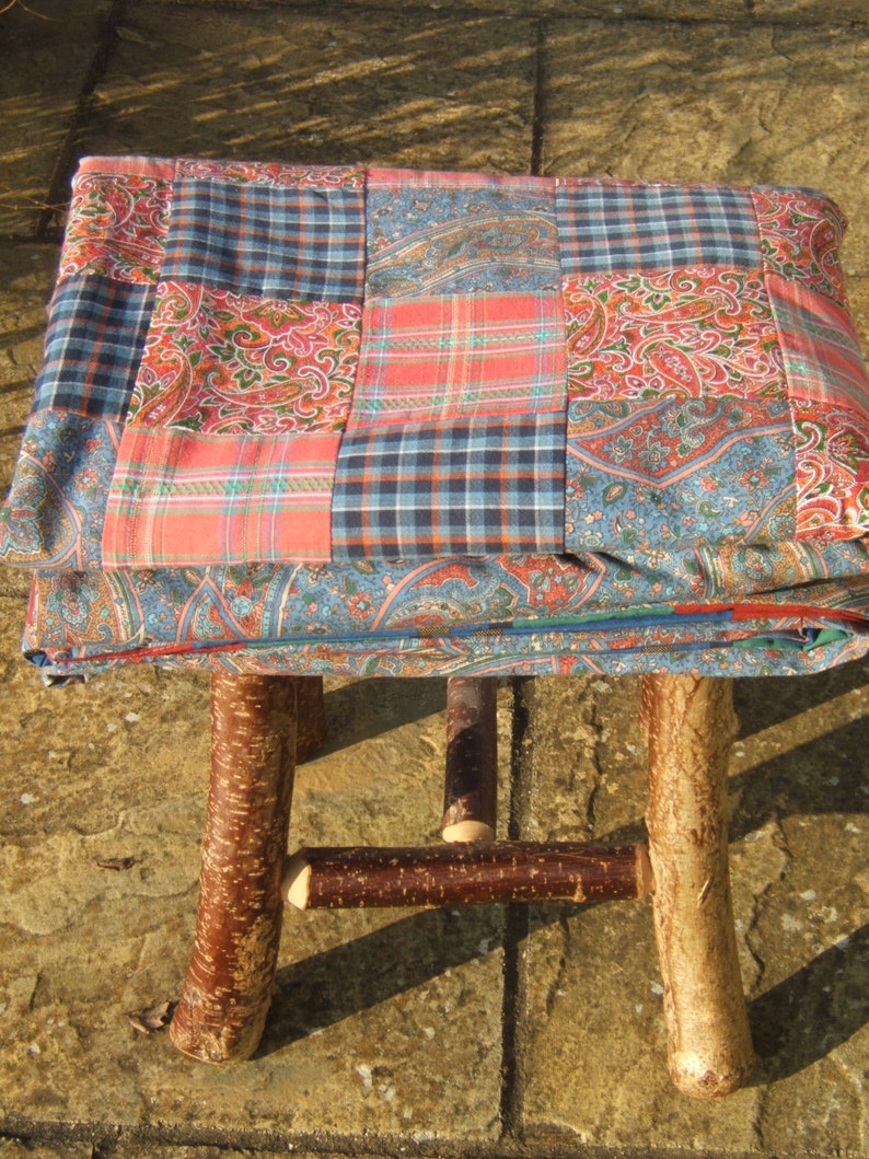 May include: A patchwork quilt made of blue, red, and white fabric is draped over a wooden stool with a natural bark finish. The stool has three legs and a simple design.