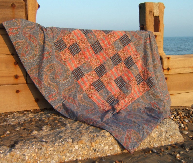 May include: A patchwork quilt with a blue paisley pattern on the border and a red and black plaid pattern in the center. The quilt is folded and draped over a wooden surface.