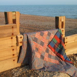 May include: A patchwork quilt with a blue, red, and green plaid pattern is draped over a wooden fence post on a beach. The quilt is folded in half and the plaid pattern is visible on the top half of the quilt.