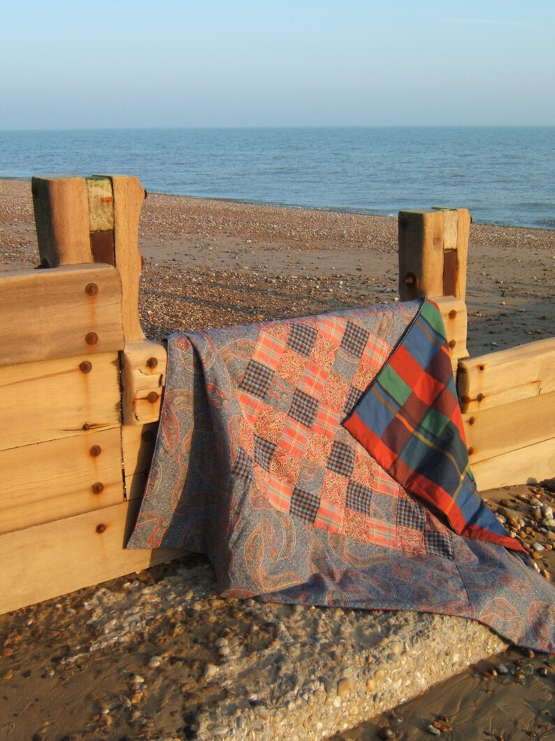 May include: A patchwork quilt with a blue, red, and green plaid pattern is draped over a wooden fence post on a beach. The quilt is folded in half and the plaid pattern is visible on the top half of the quilt.