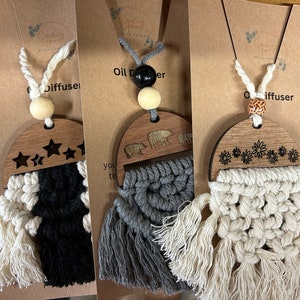 May include: Three wooden car air fresheners with macrame tassels. The first has a black and white tassel and stars carved into the wood. The second has a gray and white tassel and bears carved into the wood. The third has a white tassel and flowers carved into the wood. Each has a wooden bead and a black cord.