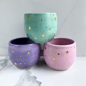 May include: Three ceramic planters in different pastel colours with gold star accents. The planters are stacked on top of each other, with the green planter on top, the purple planter in the middle, and the pink planter on the bottom.