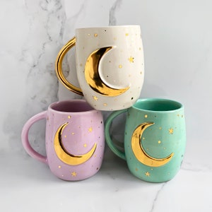 May include: Three ceramic mugs in cream, lavender, and mint green, each featuring a gold crescent moon and star design. The cream mug has a gold handle. The mugs are decorated with gold stars and dots.