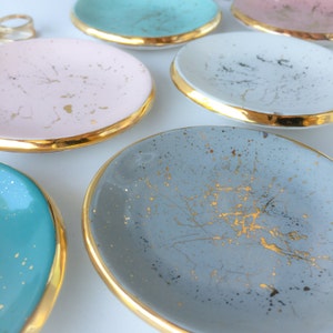 Puede incluir: Conjunto de cinco platos decorativos pequeños con bordes dorados. Los platos son de varios colores, incluyendo azul claro, rosa, gris y blanco. Cada plato tiene un patrón moteado con detalles dorados.