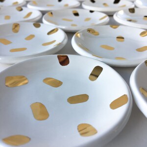 Puede incluir: Un juego de platos de anillo de cer&aacute;mica blanca con un patr&oacute;n de confeti dorado. Los platos est&aacute;n dispuestos en un patr&oacute;n escalonado.