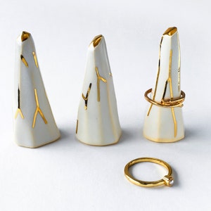 May include: Three white ceramic ring holders with gold accents. The holders are shaped like small mountains and have a textured surface. A gold ring with a small pearl is also shown.