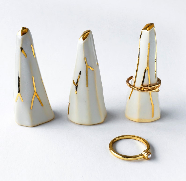 May include: Three white ceramic ring holders with gold accents. The holders are shaped like small mountains and have a textured surface. A gold ring with a small pearl is also shown.