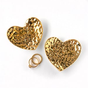 Puede incluir: Dos platos dorados en forma de coraz&oacute;n con textura martillada. Los platos est&aacute;n sobre un fondo blanco y hay dos anillos dorados en el centro de la imagen.