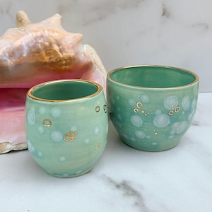 May include: Two green ceramic cups with gold rims and white and gold speckles. The cups are on a white marble surface.