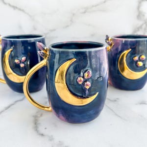 May include: Three ceramic mugs with a crescent moon design in gold, set against a dark blue background with pink and purple accents. Each mug has a crystal-like handle.
