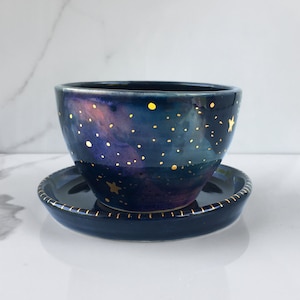 May include: A ceramic bowl and saucer set with a celestial design. The bowl is deep blue with a galaxy-like pattern in purple and gold. Gold dots and stars are scattered across the surface. The saucer is dark blue with a gold rim.