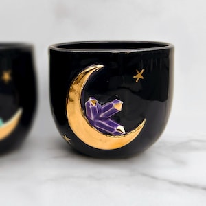 Könnte beinhalten: Schwarze Keramik-Tasse mit goldenem Halbmond und lila Kristallcluster-Design. Goldene Sterne sind um den Mond verstreut. Die Tasse hat eine abgerundete Form.