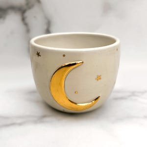 Könnte beinhalten: Eine weiße Keramiktasse mit einem goldenen Halbmond- und Sternenmuster.
