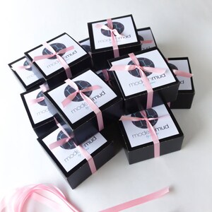 Puede incluir: Una pila de cajas de regalo negras con una cinta rosa atada alrededor de cada caja. Las cajas est&aacute;n etiquetadas con "mode mud by" en texto blanco.