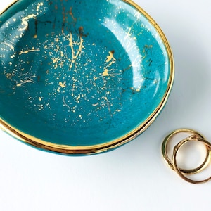 May include: A teal blue ceramic ring dish with a gold rim and gold speckles. The dish is sitting on a white surface.