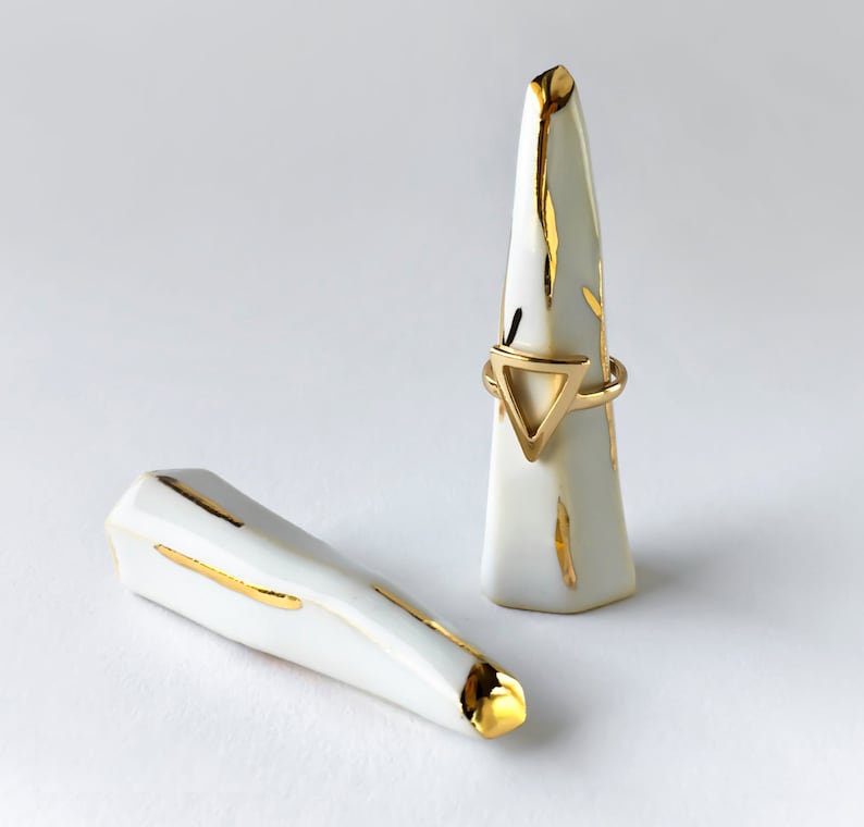 May include: Two white ceramic ring holders with gold accents. One holder is a cone shape and the other is a long, thin shape. A gold ring with a triangle design is on the cone-shaped holder.
