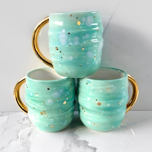 Op de afbeelding: Drie turquoise keramische mokken met gouden handvatten zijn op elkaar gestapeld. De mokken hebben een getextureerd, golvend ontwerp en zijn bespikkeld met gouden en witte accenten. De mokken zijn in een driehoekige formatie gerangschikt.