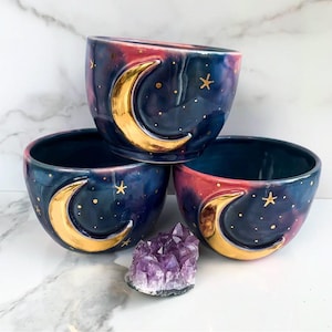 Può includere: Tre ciotole in ceramica con un design a galassia. Le ciotole sono blu e rosa con lune crescenti dorate e stelle. Le ciotole sono impilate l'una sull'altra.