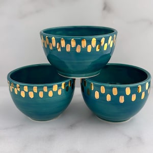 May include: Three teal ceramic bowls with gold accents. The bowls are stacked, showcasing their rounded shape and the hand-painted gold details. The bowls are a vibrant teal color.