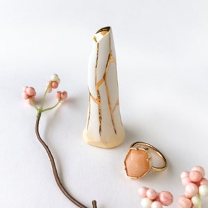May include: A white ceramic vase with gold crackle glaze and a small, peach-coloured ring with a gold setting.