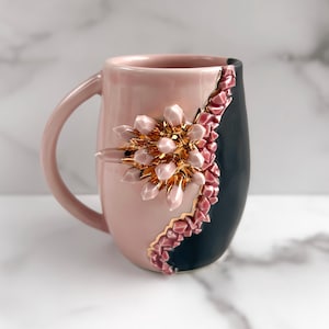 May include: A pink ceramic mug with a black section and a decorative floral design. The mug features a pink handle and a gold-colored floral embellishment with pink crystal-like accents. The mug is a unique piece of drinkware.