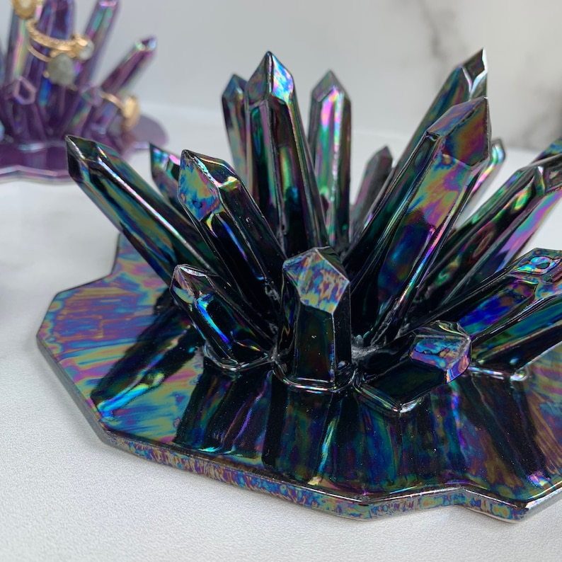 May include: Iridescent black crystal-shaped jewelry holder. The holder has a cluster of pointed crystal formations on a hexagonal base. The surface reflects a rainbow of colors. A second purple holder is visible in the background.