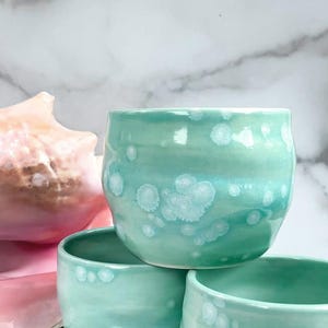 May include: Stack of three teal ceramic mugs with white speckles. The mugs are stacked on a white marble surface.