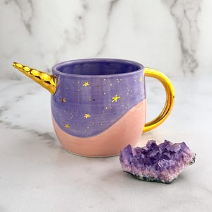 Puede incluir: Una taza de cerámica morada y rosa con un asa de cuerno de unicornio dorado y estrellas doradas. La taza está sobre una superficie de mármol blanco junto a un cristal de amatista morado.